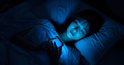Person lying in bed at night scrolling smartphone with blue light glow illuminating face in dark bedroom