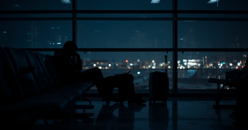 Exhausted traveller sitting alone in dark airport terminal at night with city lights visible through window